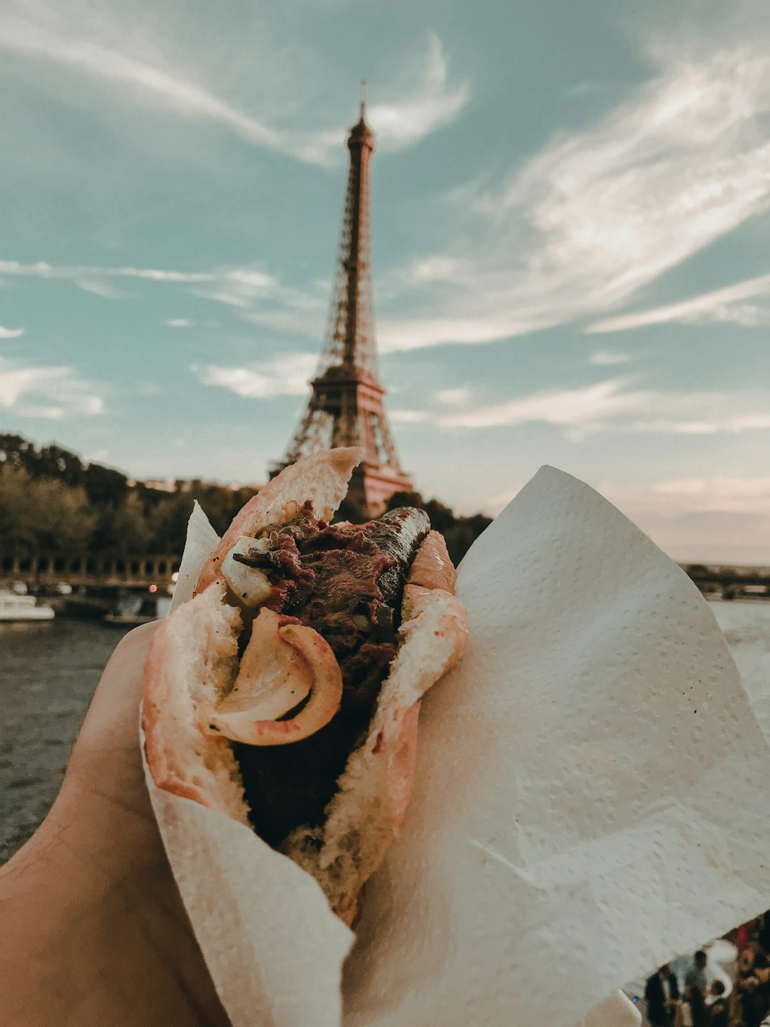 Hand holding a halal pastrami sandwich wrapped in paper with the Eiffel Tower and Seine River at dusk