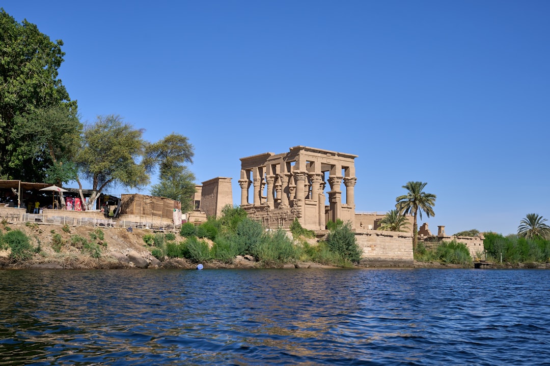 Philae Temple kiosk with ancient columns rising above the Nile, surrounded by palms and green shrubs under a clear blue sky