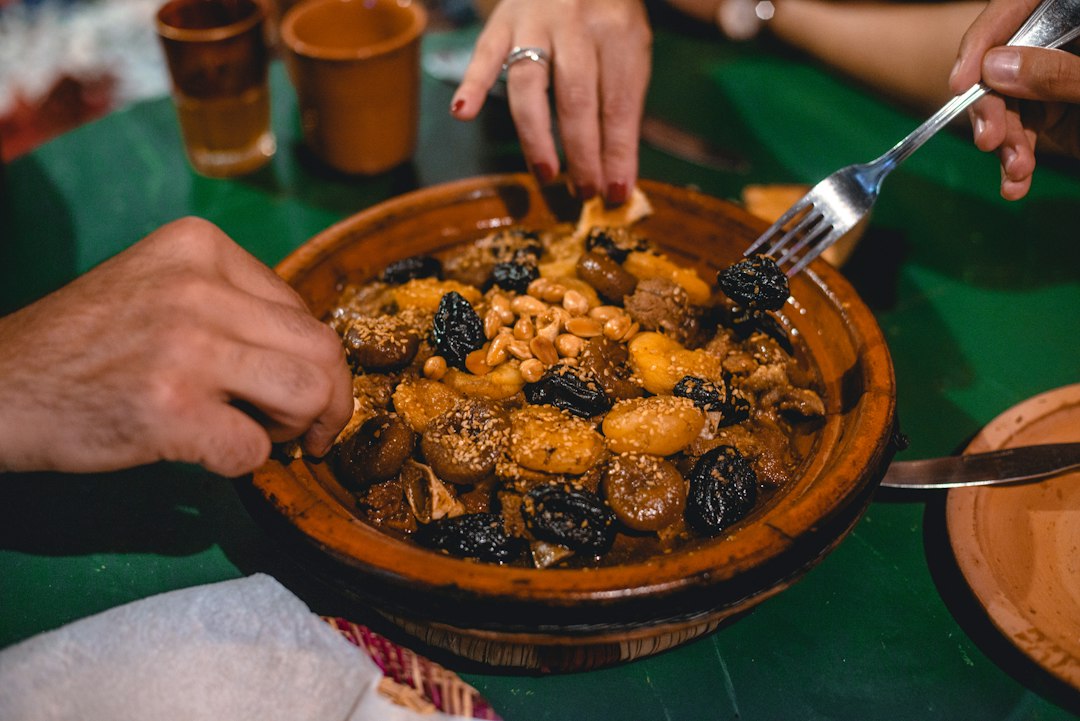 Hands sharing a Moroccan clay tagine filled with stewed meat, prunes, almonds, and sesame seeds on a green table