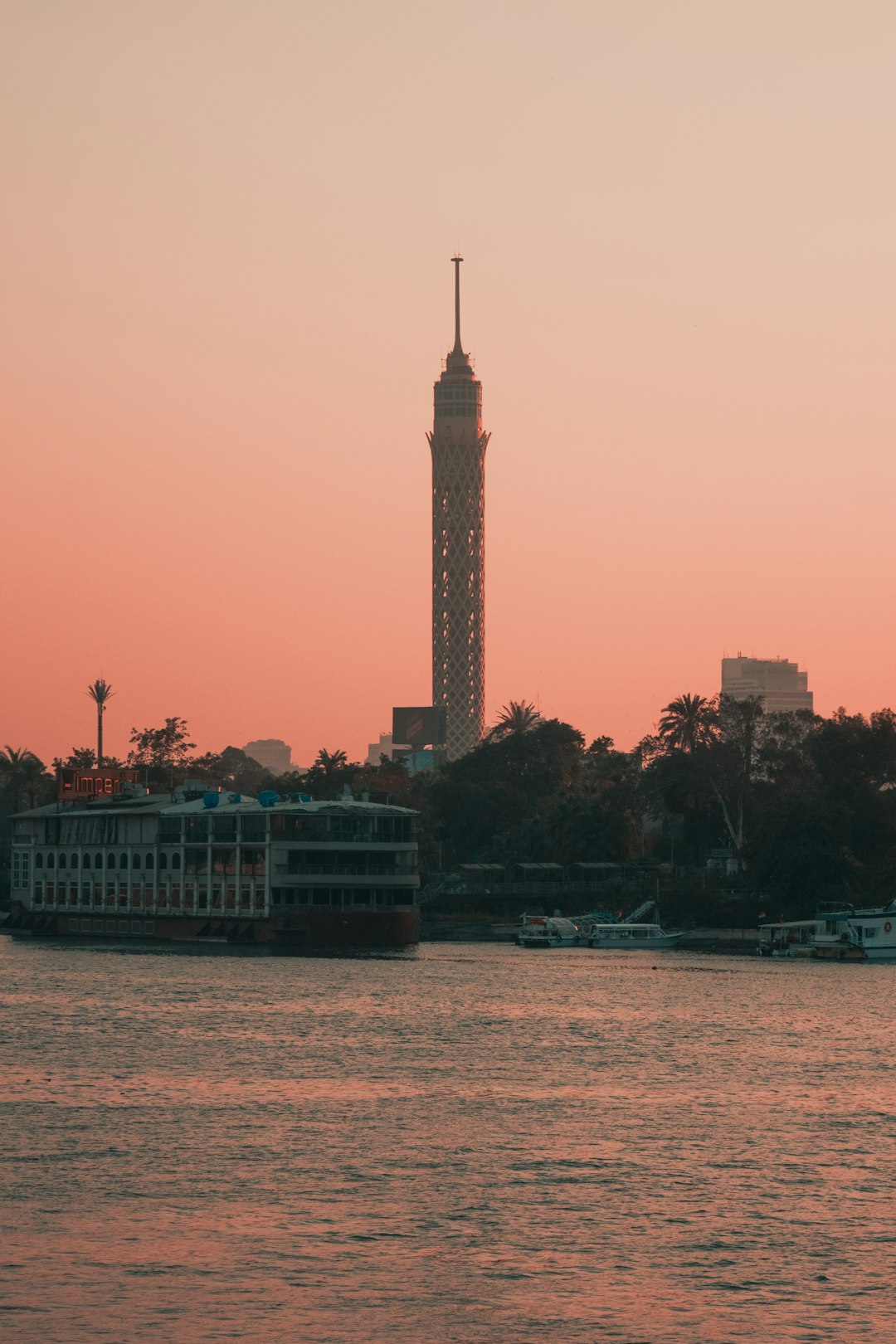 Cairo Tower rising above the Nile at sunset with moored riverboats and a pink-orange sky