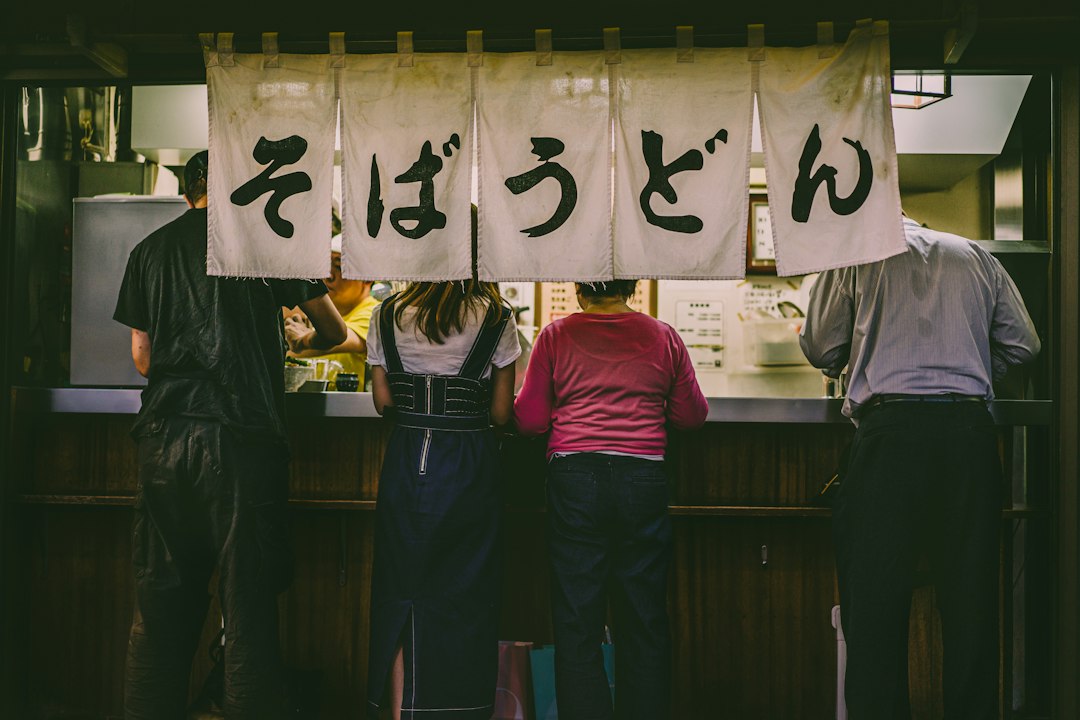 Four diners stand at a Japanese counter restaurant beneath a noren curtain reading soba and udon in kanji