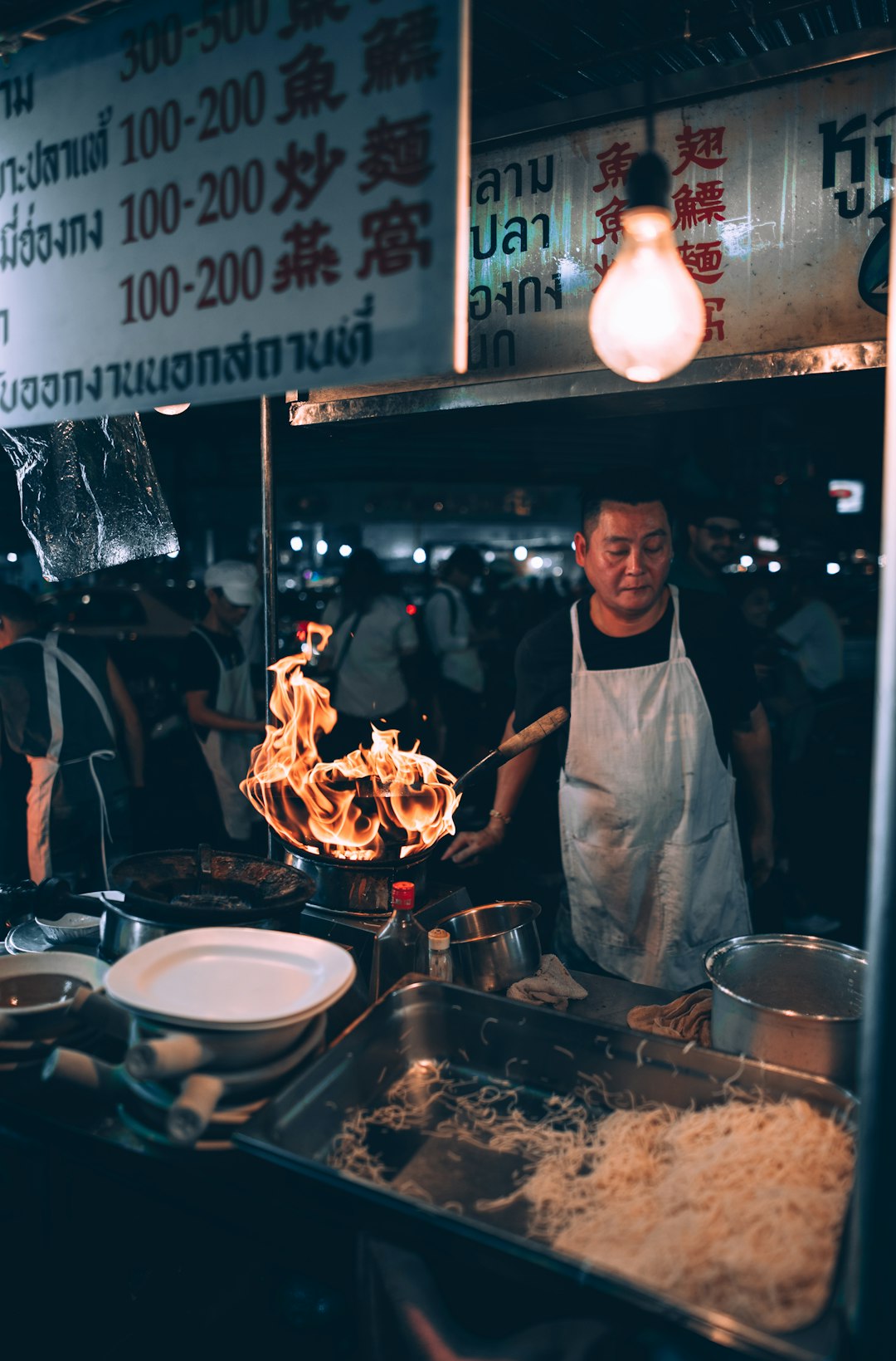 Street food chef in white apron stir-frying with dramatic wok flames at a Bangkok night market stall
