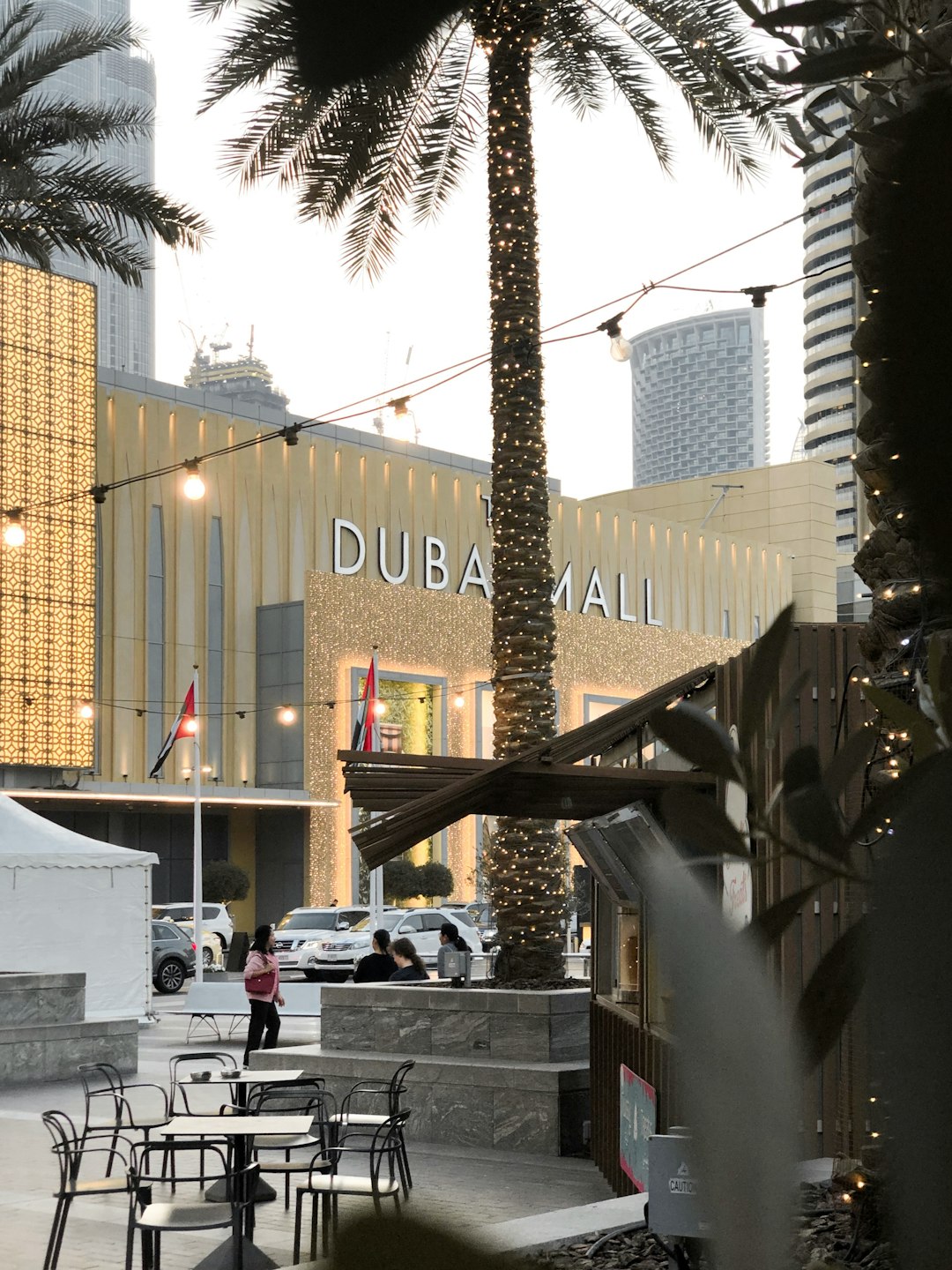 Outdoor café seating with fairy-lit palm trees in front of The Dubai Mall entrance, skyscrapers in background