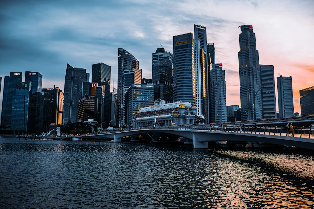 Singapore CBD skyline at dusk with glowing skyscrapers reflected in Marina Bay and a pedestrian bridge in the foreground