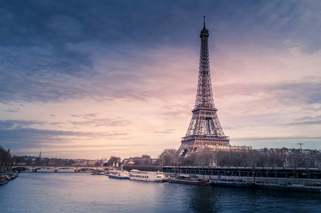 Eiffel Tower rising above the Seine River at dusk, with moody purple skies and riverboats docked along the Paris bank