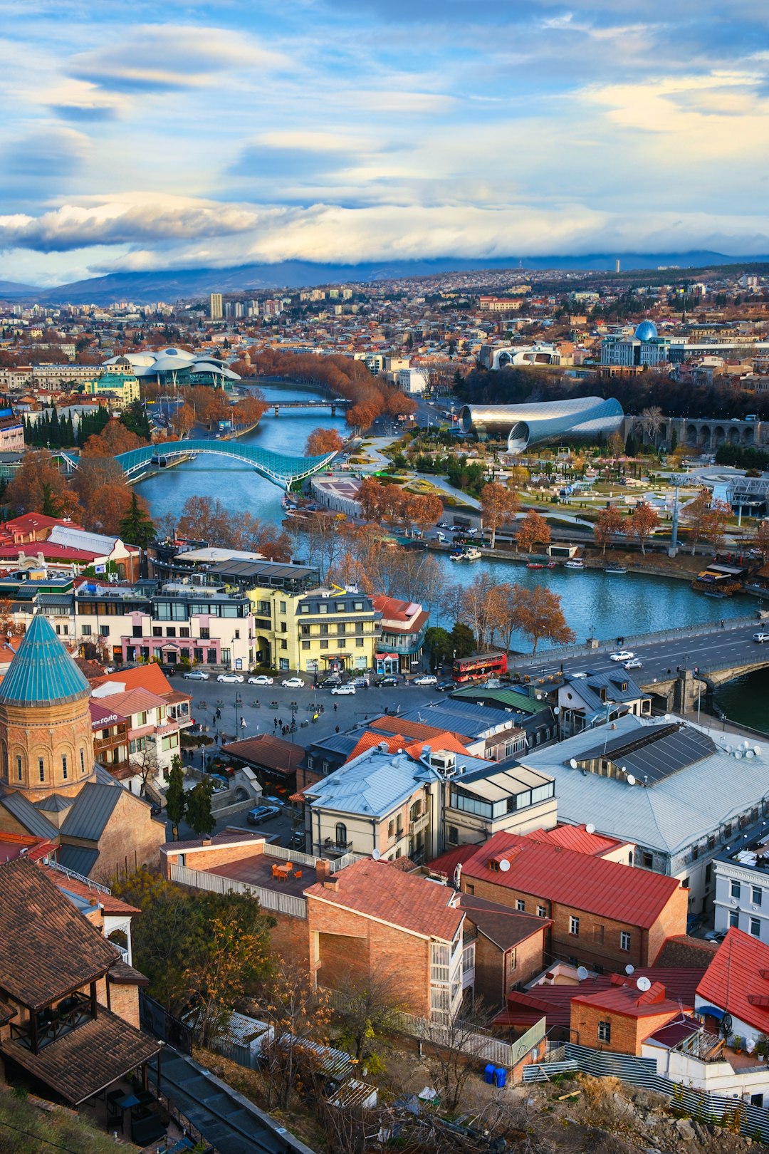 Aerial view of Tbilisi Old Town, Mtkvari River, Peace Bridge, and modern landmarks under a cloudy sky, Georgia